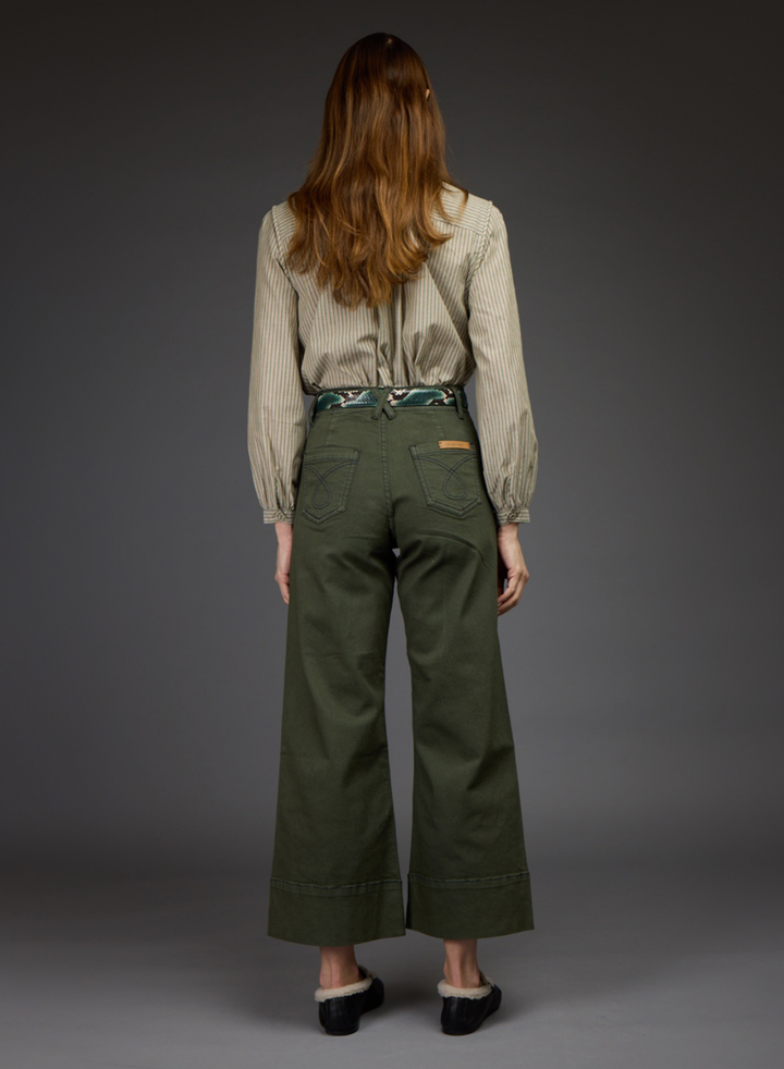 A woman viewed from the back wearing olive green wide-leg cropped pants, a beige ribbed long-sleeve blouse, and black shoes, standing against a dark gray studio background.