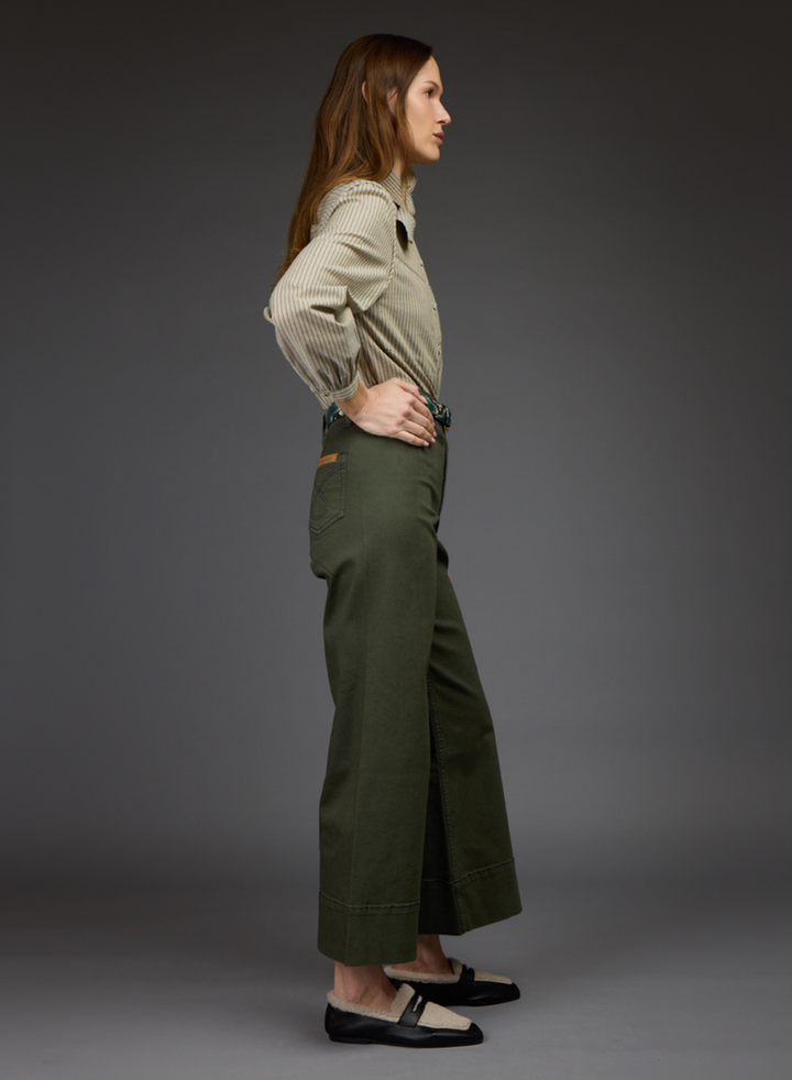 A woman stands in profile against a dark gray studio background, wearing olive green wide-leg cropped pants, a light beige ribbed long-sleeve shirt, and black-and-beige flat shoes.
