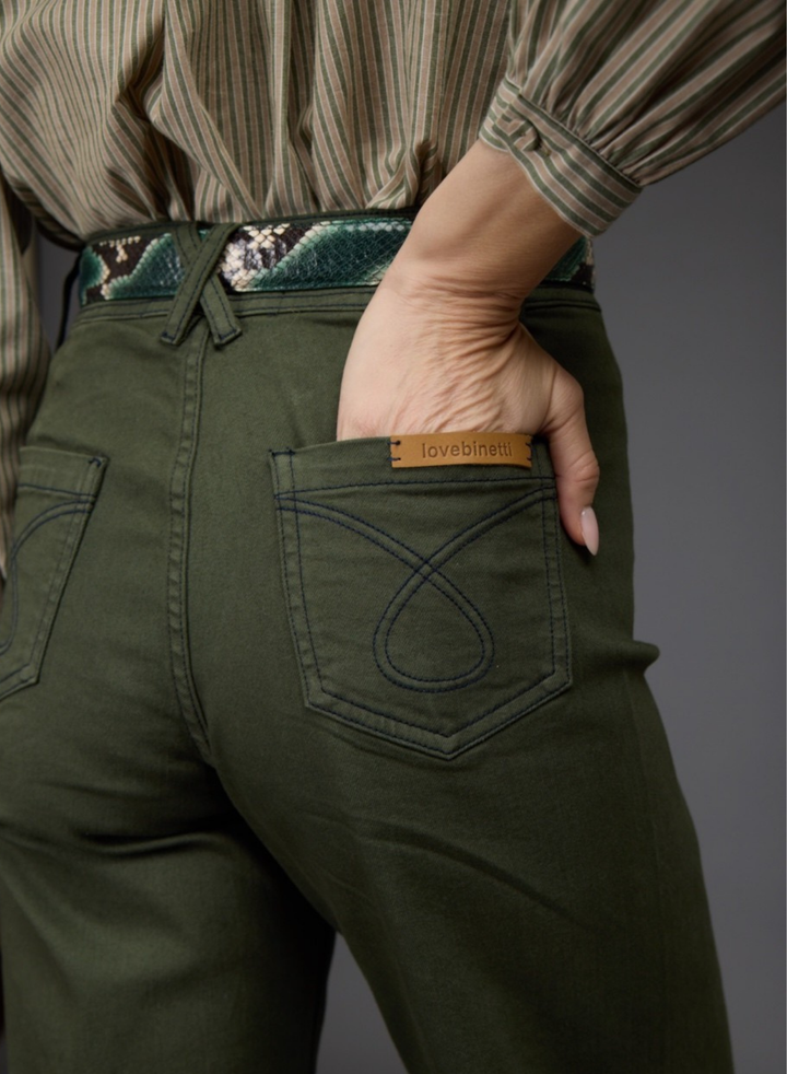 Close-up back view of a person wearing olive green pants with a decorative patterned belt and a tan leather brand tab on the back pocket.