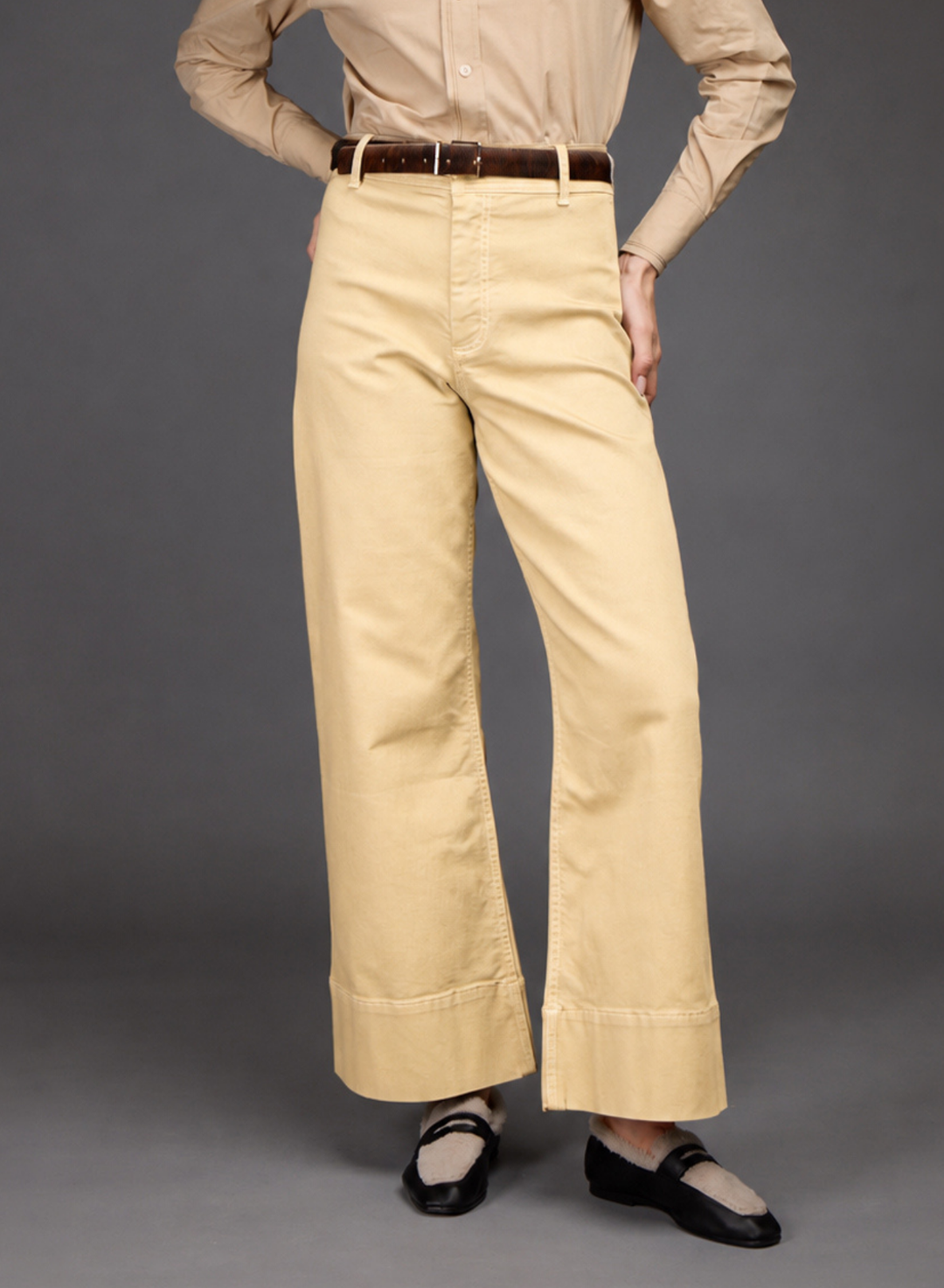 A person wearing light beige wide-leg cropped pants with a brown belt, paired with a beige shirt and black flats, photographed against a plain gray studio background.