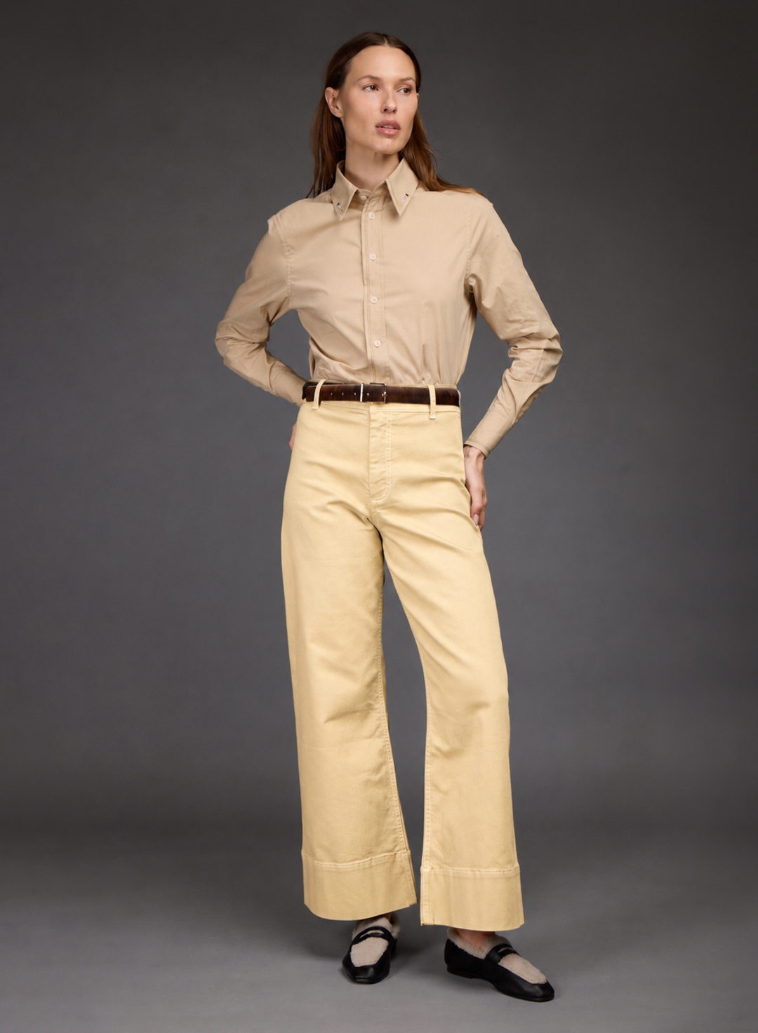 A woman modeling a beige long-sleeve button-up shirt and light beige wide-leg trousers with a brown belt, paired with black Mary Jane shoes, against a dark gray studio background.