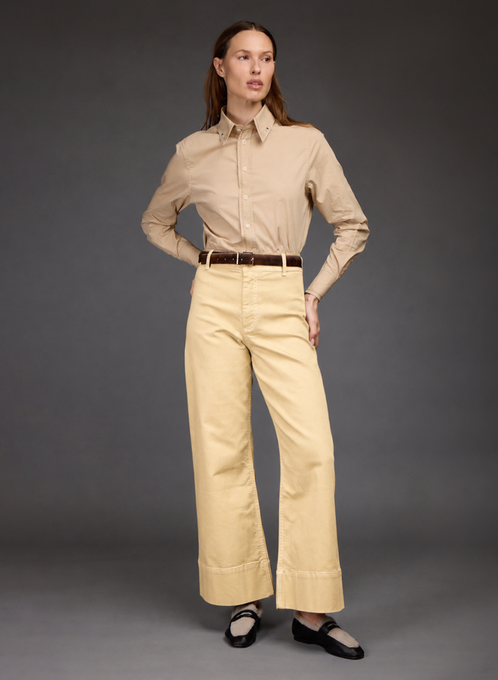 A woman modeling a beige long-sleeve button-up shirt and light beige wide-leg trousers with a brown belt, paired with black Mary Jane shoes, against a dark gray studio background.