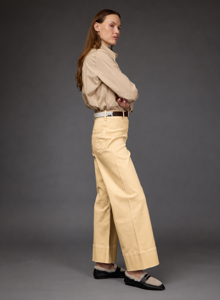 A woman stands in profile wearing a beige long-sleeve button-up shirt, light beige wide-leg cropped pants with a brown belt, and black Mary Jane flats against a dark gray studio background.