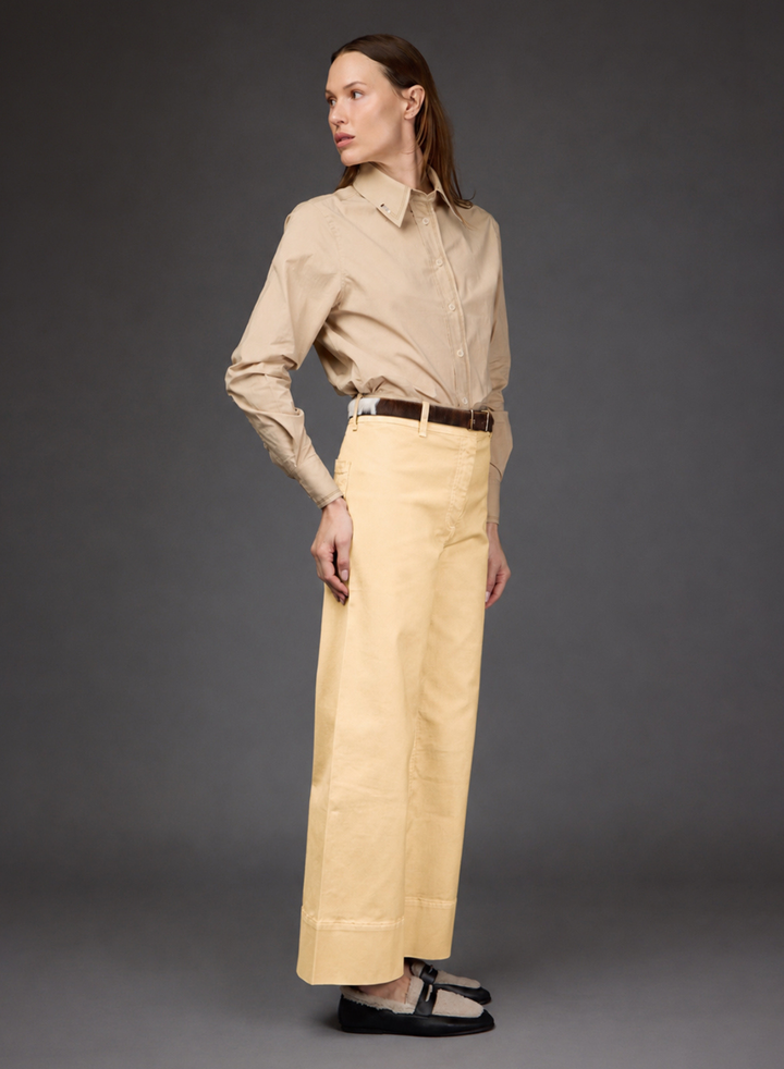 A model stands in profile wearing a beige long-sleeve button-up shirt tucked into light yellow wide-leg trousers, with a dark brown belt and black flats, against a dark gray studio background.
