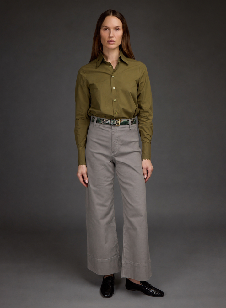 A woman stands against a dark gray studio background wearing a long-sleeve olive green button-up shirt tucked into high-waisted light gray wide-leg trousers, with a patterned belt and black loafers.