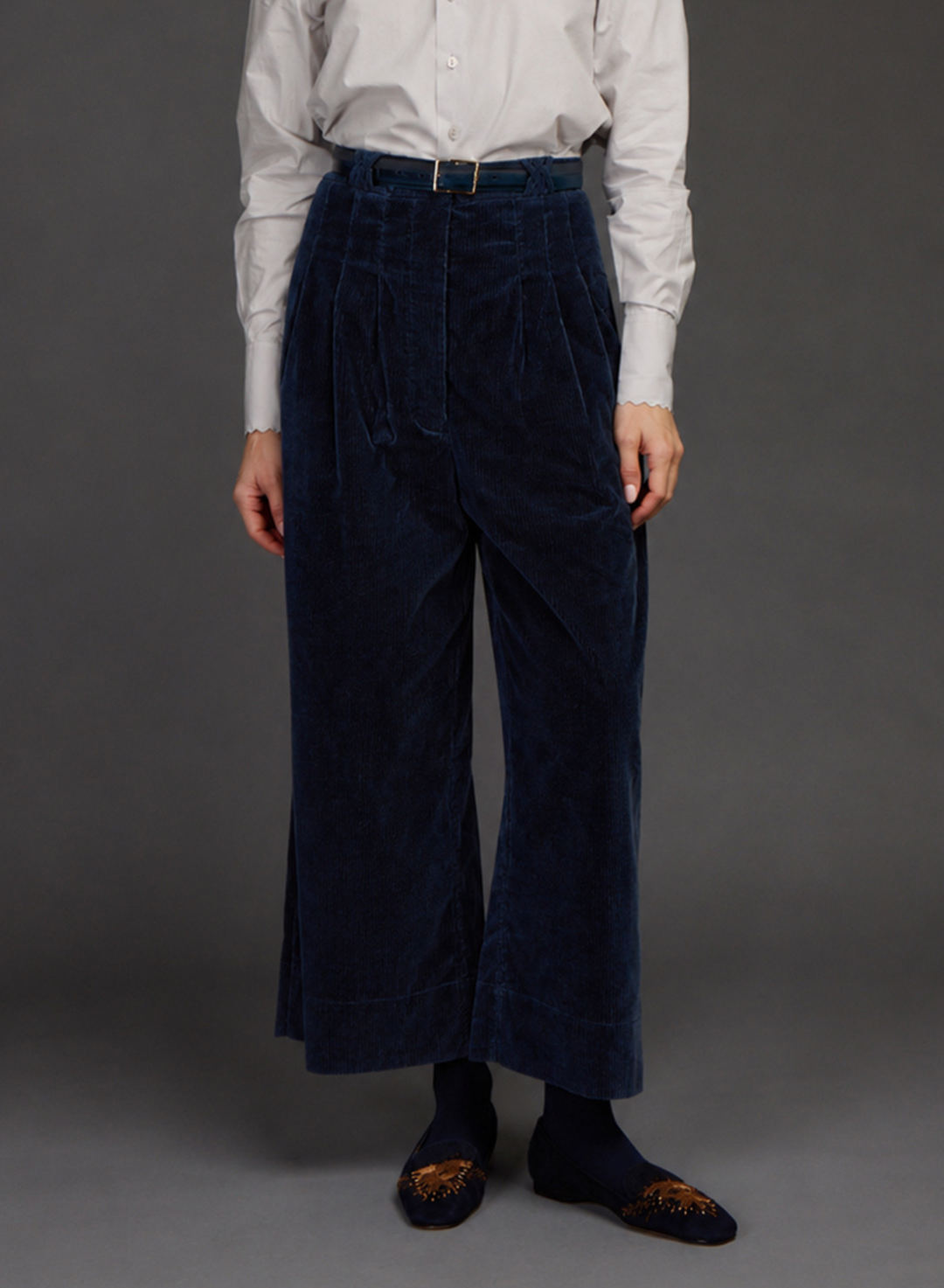 A person wearing dark navy corduroy wide-leg trousers with a matching belt, paired with a white button-down shirt by Love BInetti, dark socks, and patterned slip-on shoes, standing against a dark gray studio background.