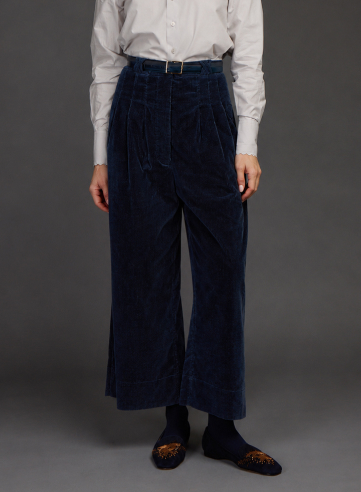 A person wearing dark navy corduroy wide-leg trousers with a matching belt, paired with a white button-down shirt by Love BInetti, dark socks, and patterned slip-on shoes, standing against a dark gray studio background.