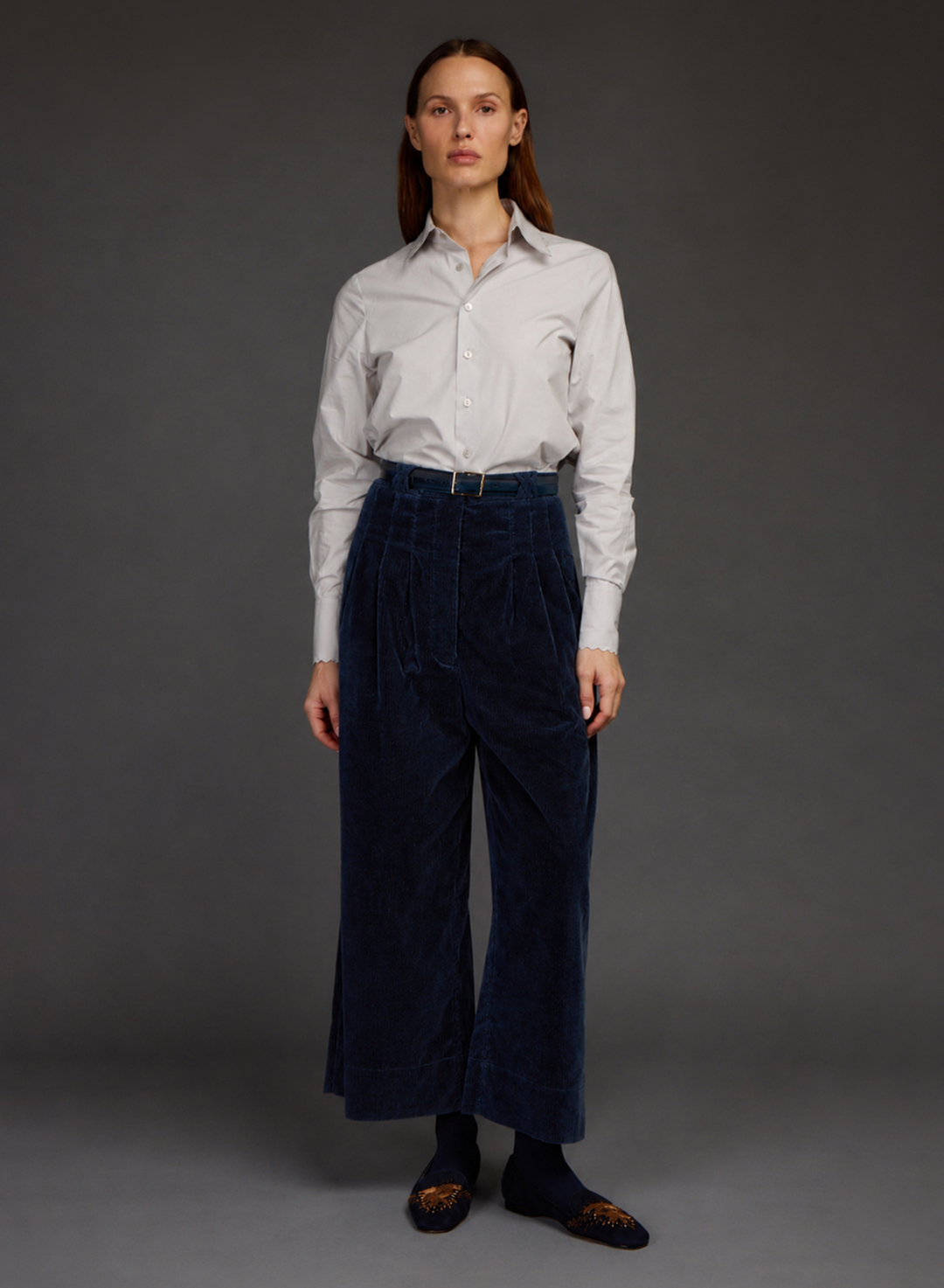 A model stands against a dark gray studio background wearing a white long-sleeve button-down shirt tucked into high-waisted, wide-leg navy corduroy trousers with a matching belt. All by Love Binetti.