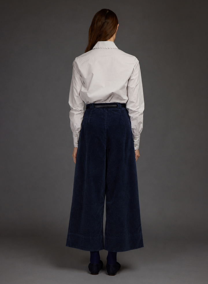A woman is shown from the back wearing a white long-sleeve button-up shirt tucked into dark navy wide-leg corduroy trousers by Love Binetti, paired with dark socks and black shoes, against a solid dark gray studio background.