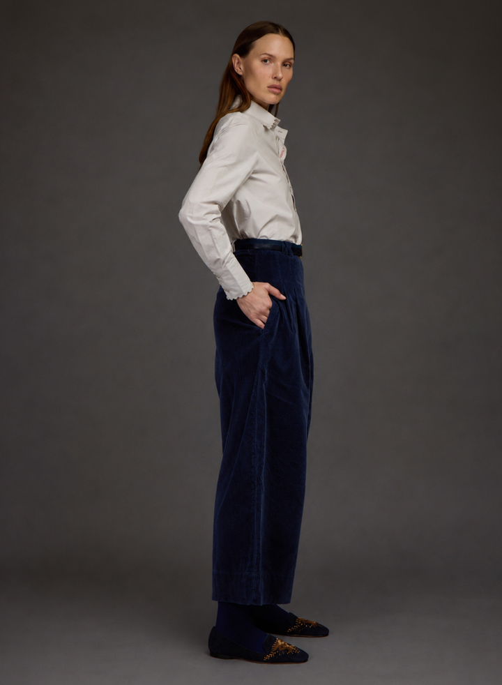 A woman stands in profile wearing a white long-sleeve button-up shirt, dark navy wide-leg trousers by Love Binetti, and dark loafers with gold embroidery, against a solid dark gray studio background.