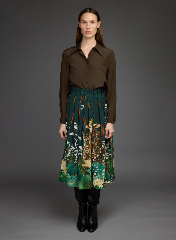 A model stands against a dark gray studio background wearing a dark brown button-up shirt, a green and beige woodland print midi skirt, and black ankle boots.