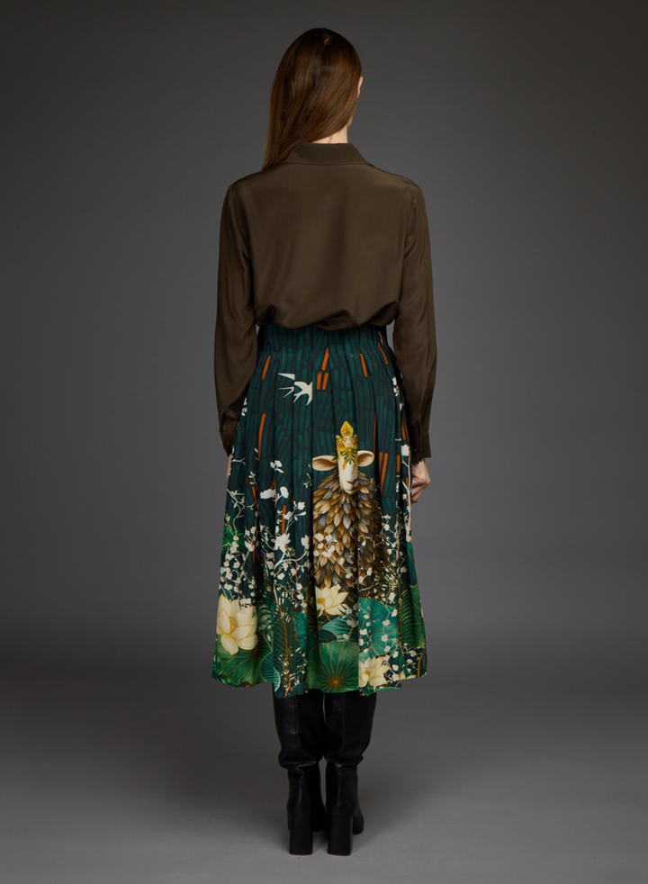 A model viewed from the back wearing a dark brown long-sleeve shirt and a pleated midi skirt with a detailed nature print featuring a deer, birds, and flowers in green, brown, and cream. She is wearing black boots against a dark gray studio background.