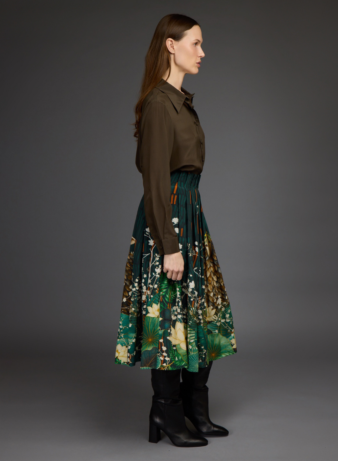 A woman stands in profile wearing a dark olive-brown button-up shirt tucked into a pleated midi skirt with a green botanical print, paired with black ankle boots, against a dark gray studio background.