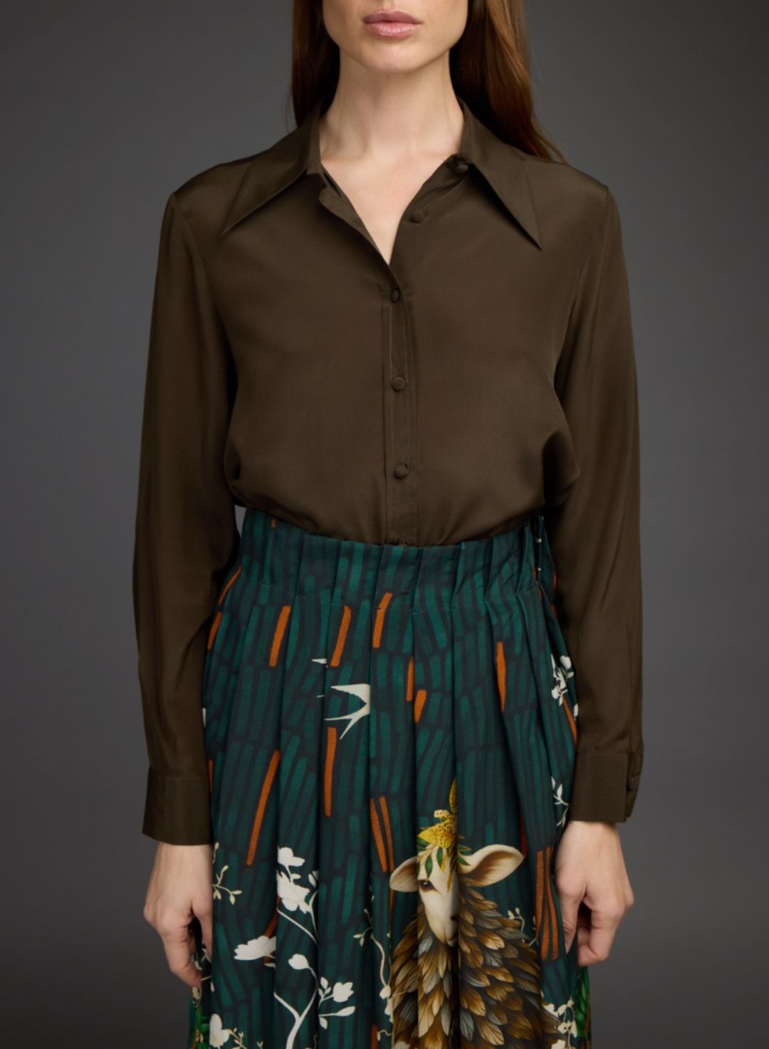 A model wearing a dark brown long-sleeve button-up shirt tucked into a dark green pleated skirt with a detailed bird and nature print, shown against a dark gray studio background.