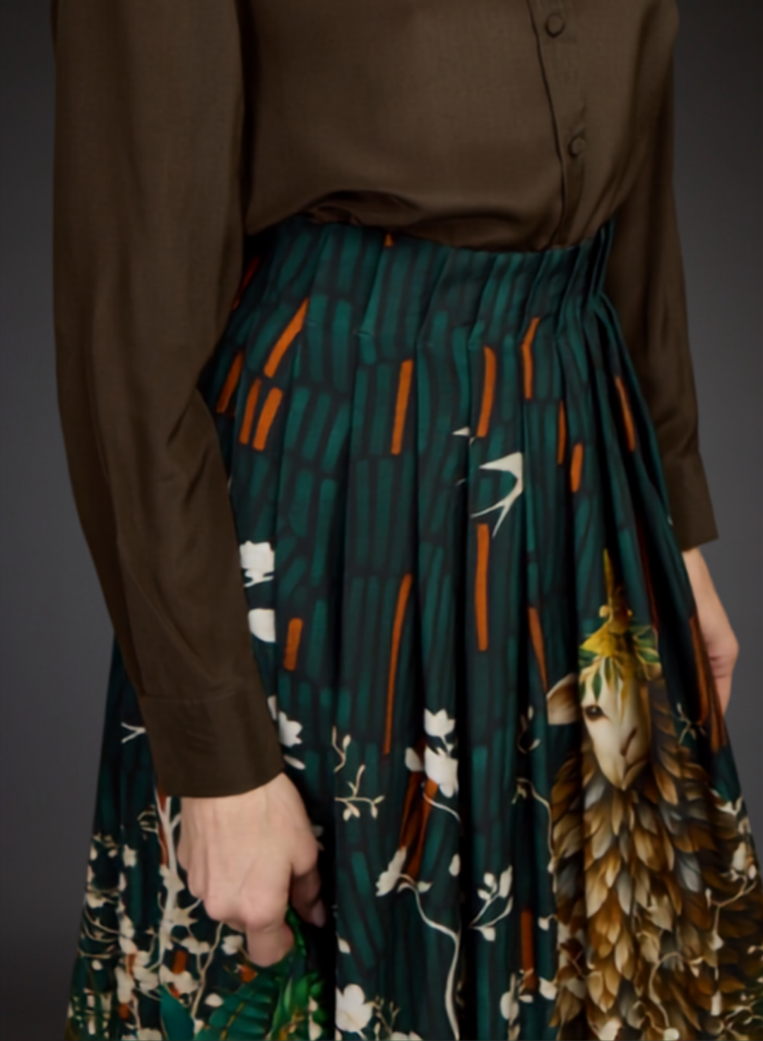A close-up of a pleated midi skirt with a dark teal green base, featuring vertical orange stripes, white floral motifs, and a large brown and gold bird illustration on the right side. The skirt is styled with a brown button-up shirt, shown on a model against a dark gray background.
