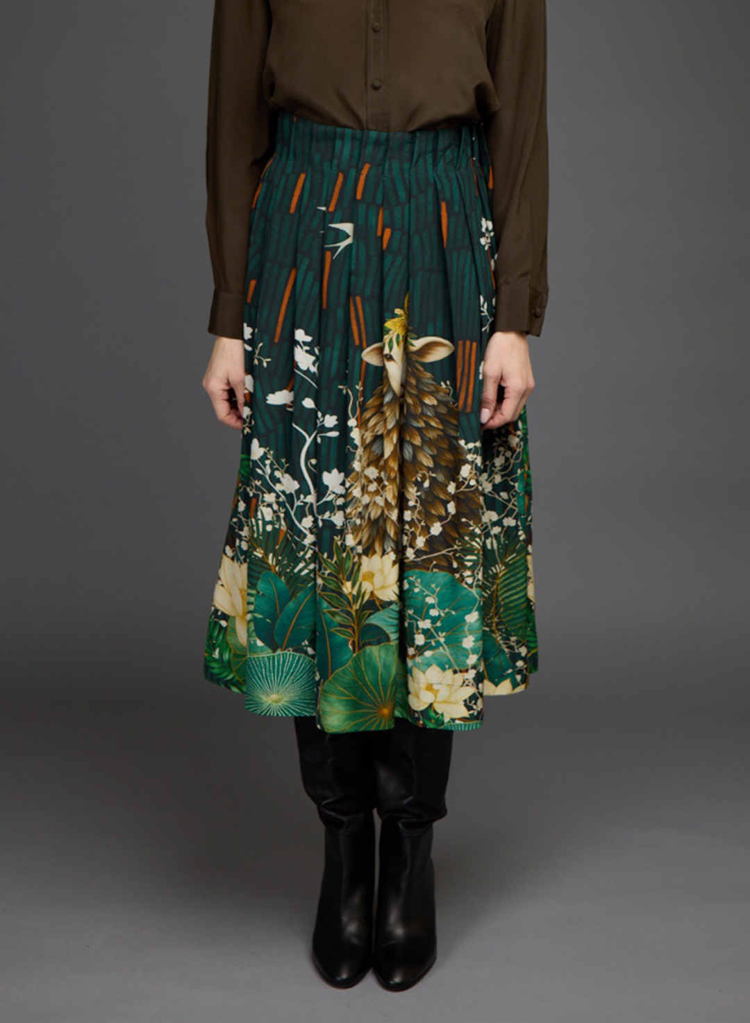 A model is shown from the chest down wearing a pleated midi skirt with a green and brown nature print featuring a deer and foliage, paired with a brown top and black boots against a gray studio background.