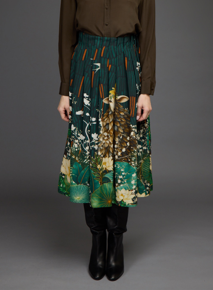 A model is shown from the chest down wearing a pleated midi skirt with a green and brown nature print featuring a deer and foliage, paired with a brown top and black boots against a gray studio background.
