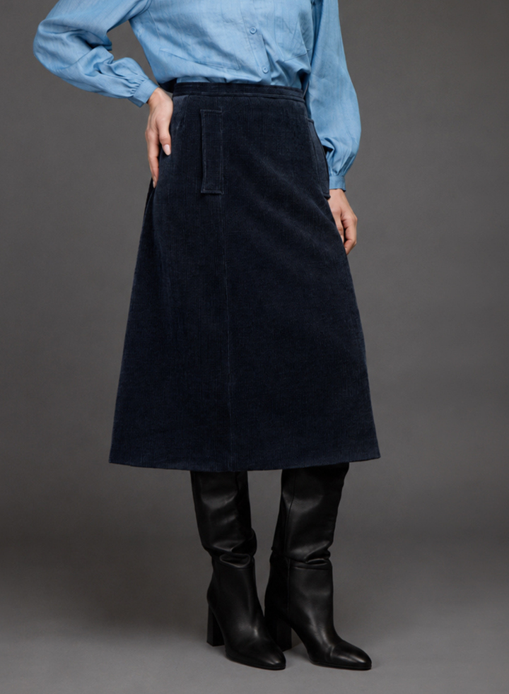 A model wearing a dark navy blue corduroy A-line skirt, paired with a light blue long-sleeve shirt and black leather knee-high boots, photographed against a neutral gray studio background.