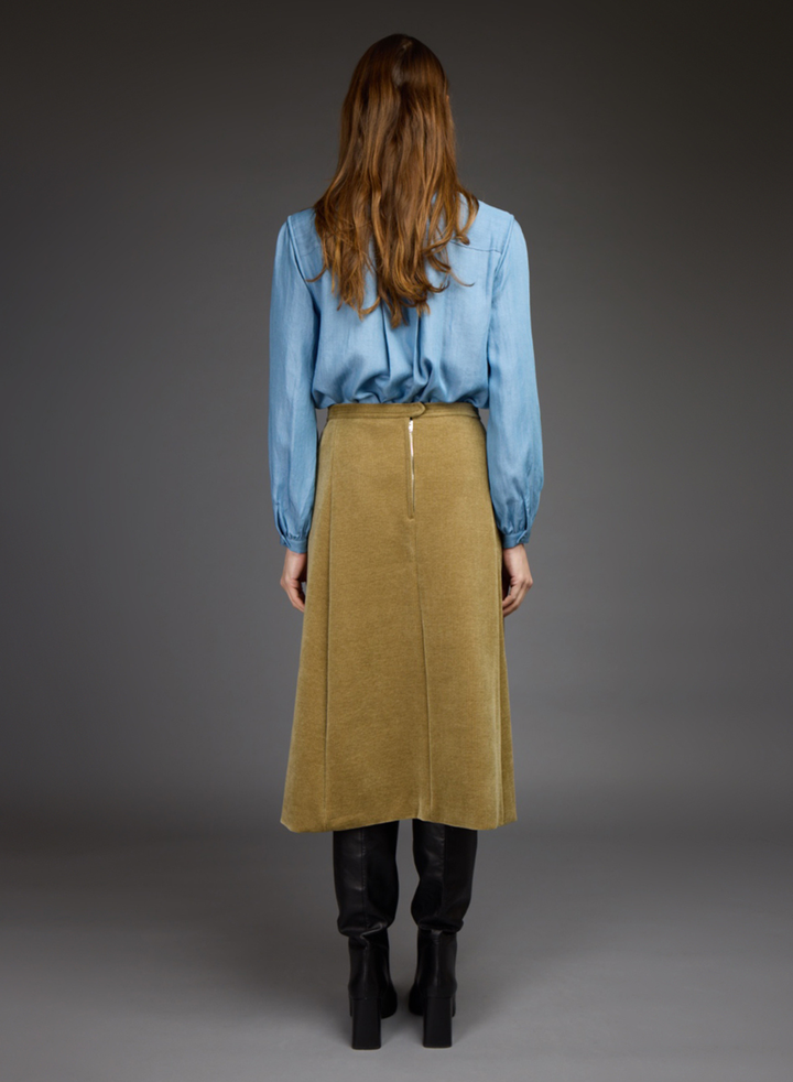A woman is shown from behind wearing a light blue long-sleeve blouse, a mustard yellow corduroy midi skirt, black leggings, and black boots, set against a dark gray studio background.