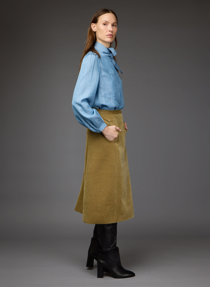 A model is shown in profile wearing a light blue long-sleeve button-up shirt, a mustard yellow corduroy skirt, and black heeled ankle boots, set against a solid dark gray studio background.