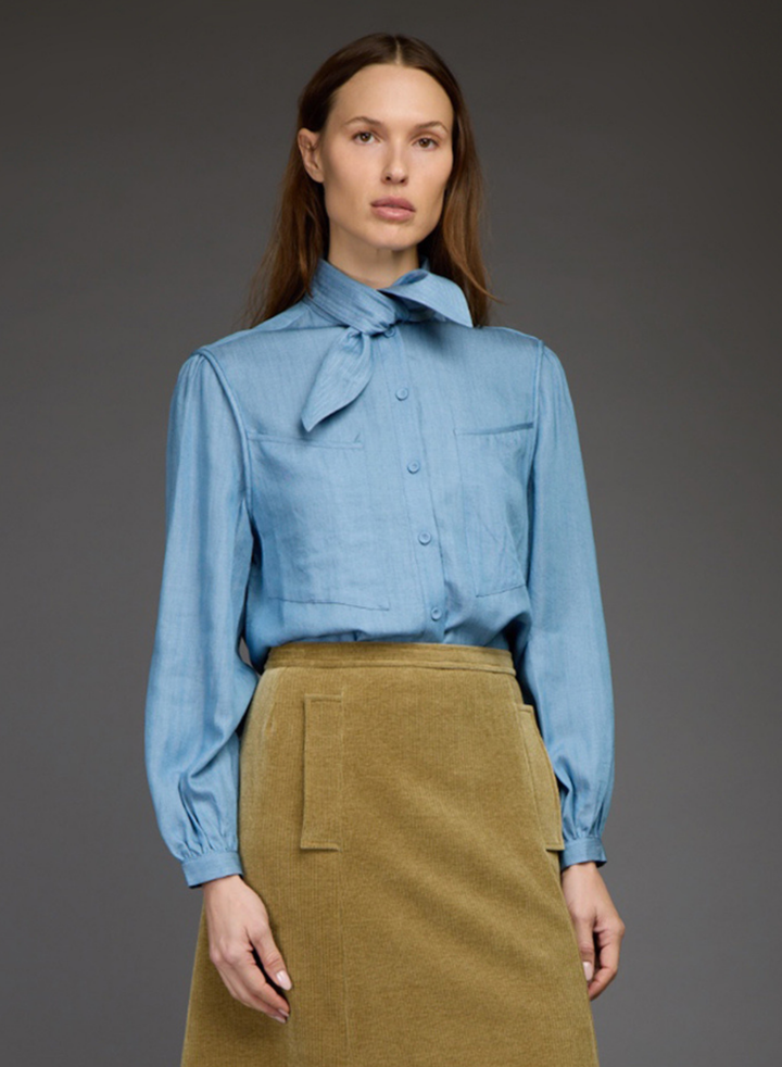 A woman modeling a light blue long-sleeve button-up shirt with a large bow tie collar, paired with a brown corduroy skirt, against a dark gray studio background.