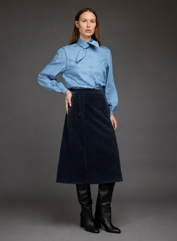 A model stands against a dark gray studio background wearing a light blue long-sleeve button-up shirt with a large bow at the collar, a dark navy corduroy A-line skirt, and black knee-high leather boots.