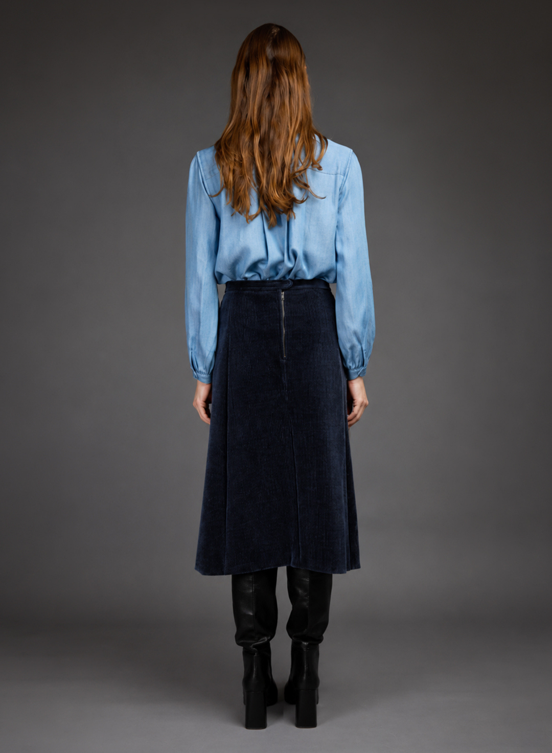 A woman is shown from the back wearing a light blue long-sleeve blouse and a dark navy blue corduroy midi skirt, paired with black knee-high boots, against a dark gray studio background.