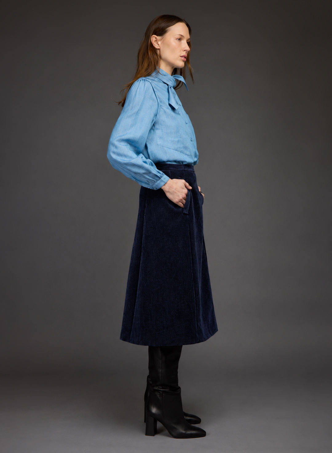 A woman stands in profile wearing a light blue long-sleeve button-up shirt, a dark navy velvet midi skirt with front pockets, black knee-high socks, and black heeled ankle boots, against a dark gray studio background.