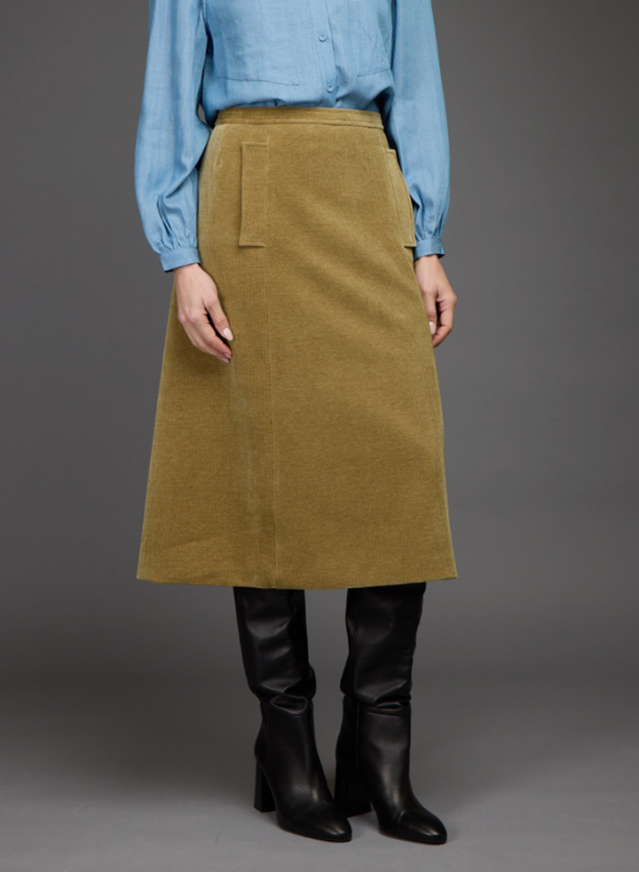 A model is shown from the waist down wearing a light blue button-up shirt, a mustard yellow corduroy A-line skirt with a side pocket, and black leather knee-high boots, set against a solid gray studio background.