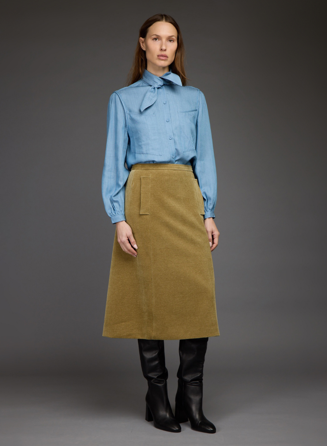A model stands against a dark gray studio background wearing a light blue long-sleeve button-up shirt with a bow tie collar, a muted olive-brown corduroy midi skirt, black leather knee-high boots, and black pointed shoes.