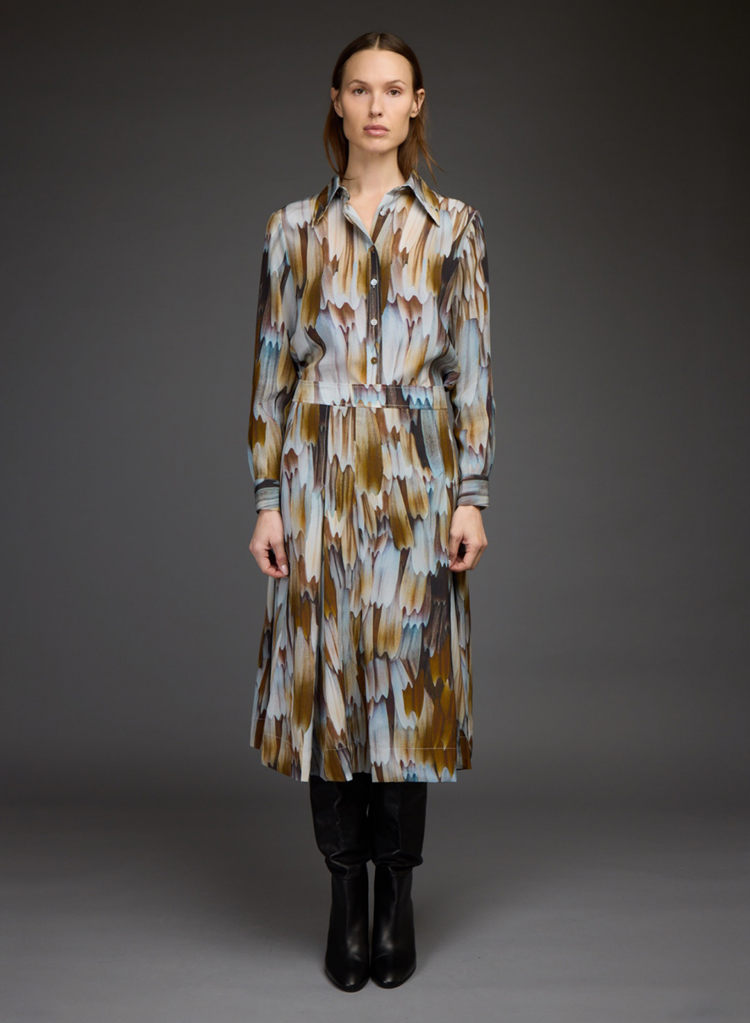 A model stands against a dark gray studio background wearing a long-sleeved, collared midi dress with an abstract brushstroke pattern in brown, beige, and light blue. She is styled with black pants and black ankle boots.