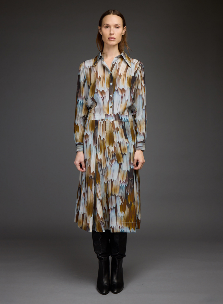 A model stands against a dark gray studio background wearing a long-sleeved, collared midi dress with an abstract brushstroke pattern in brown, beige, and light blue. She is styled with black pants and black ankle boots.