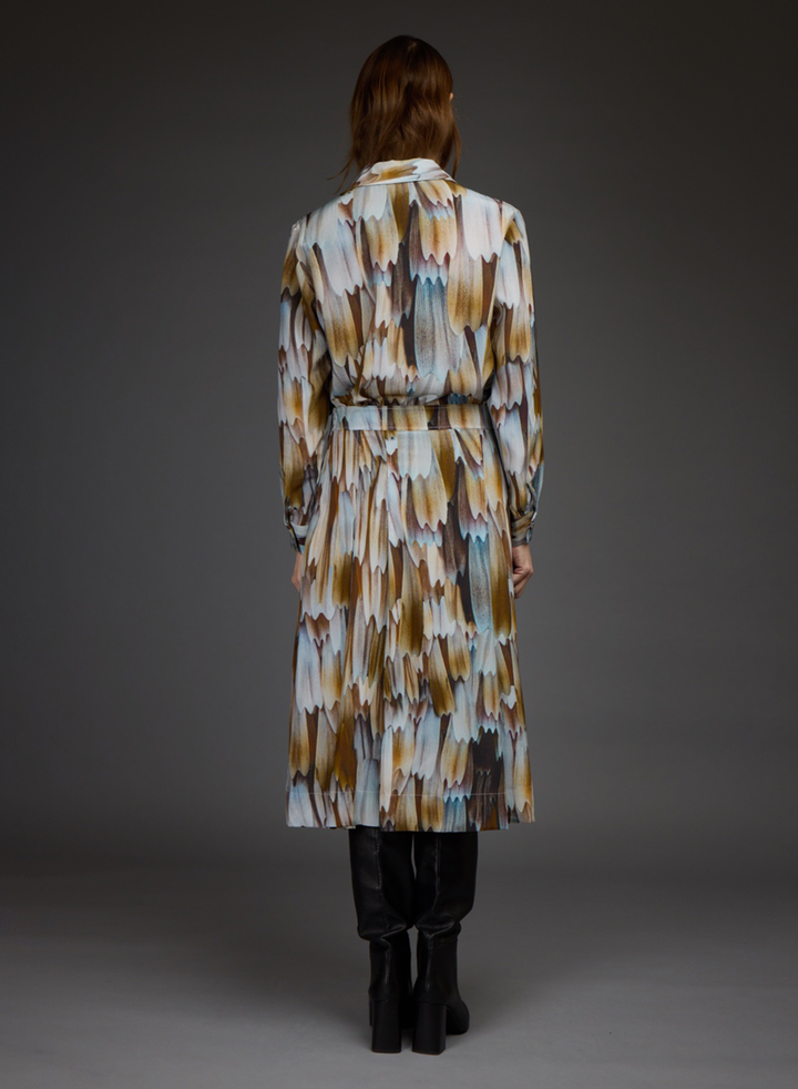 A model viewed from the back wearing a long belted coat with an abstract brushstroke pattern in beige, brown, white, and light blue, paired with black boots against a dark gray studio background.
