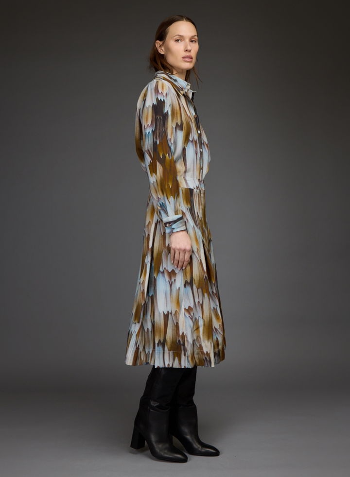 A model wearing a long-sleeved, knee-length abstract print coat in brown, beige, and light blue, paired with black leather boots, photographed against a dark gray studio background.