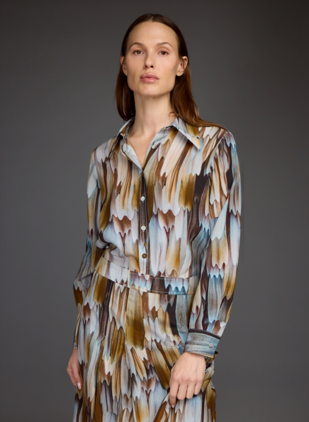 A woman modeling a long-sleeve collared shirt dress with a brown, beige, and blue abstract feather-like pattern against a dark gray studio background.