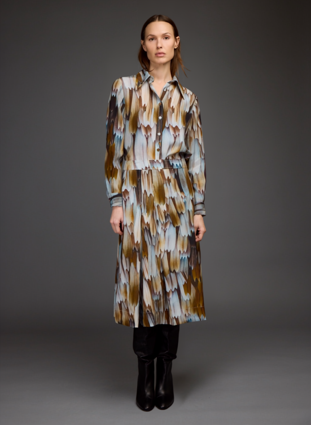 A model stands against a dark gray studio background wearing a long-sleeved button-up shirt dress with an abstract feather-like pattern in brown, beige, and light blue, paired with black boots.