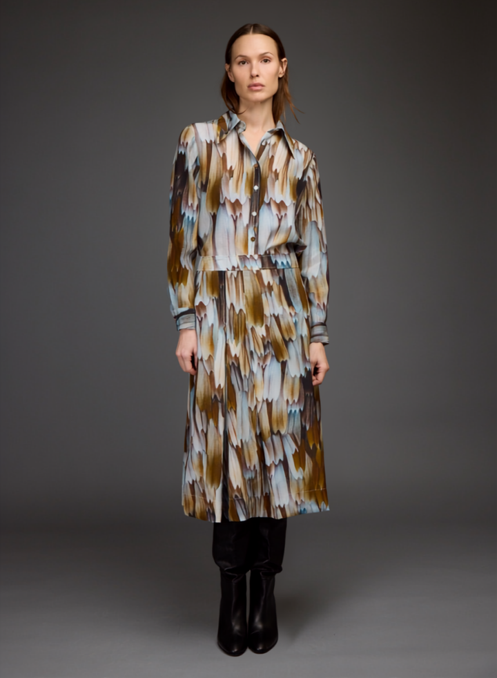 A model stands against a dark gray studio background wearing a long-sleeved button-up shirt dress with an abstract feather-like pattern in brown, beige, and light blue, paired with black boots.