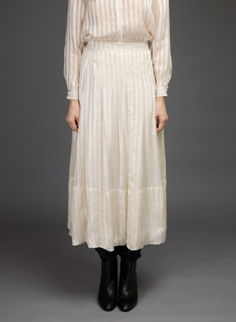 A model stands against a dark gray studio background wearing a cream-colored, long-sleeved blouse and skirt set with black leather boots.
