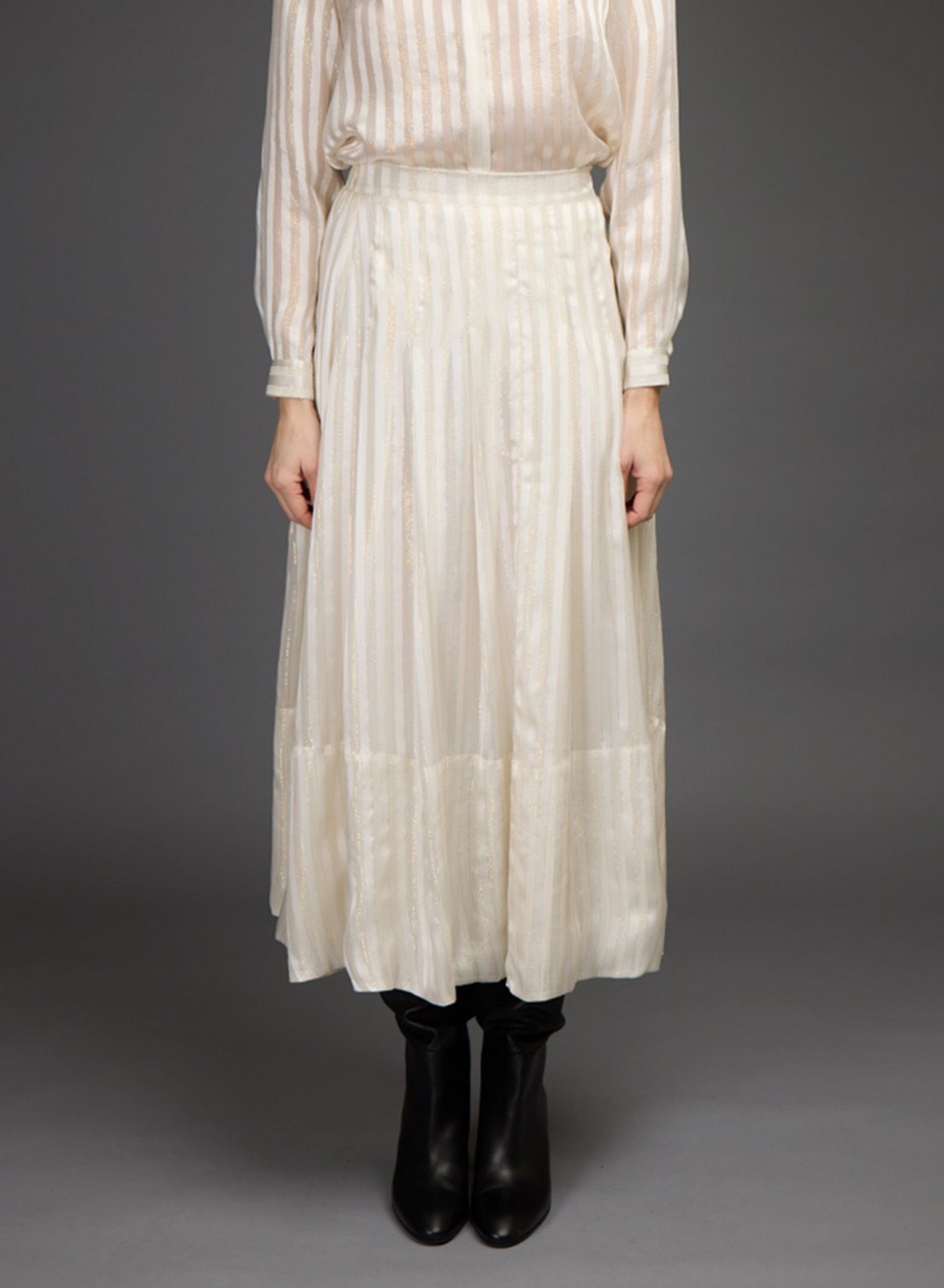 A model stands against a dark gray studio background wearing a cream-colored, long-sleeved blouse and skirt set with black leather boots.