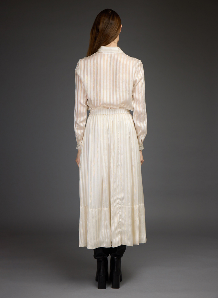 A model stands against a dark gray studio background wearing a cream-colored, long-sleeved blouse and skirt set with black leather boots.