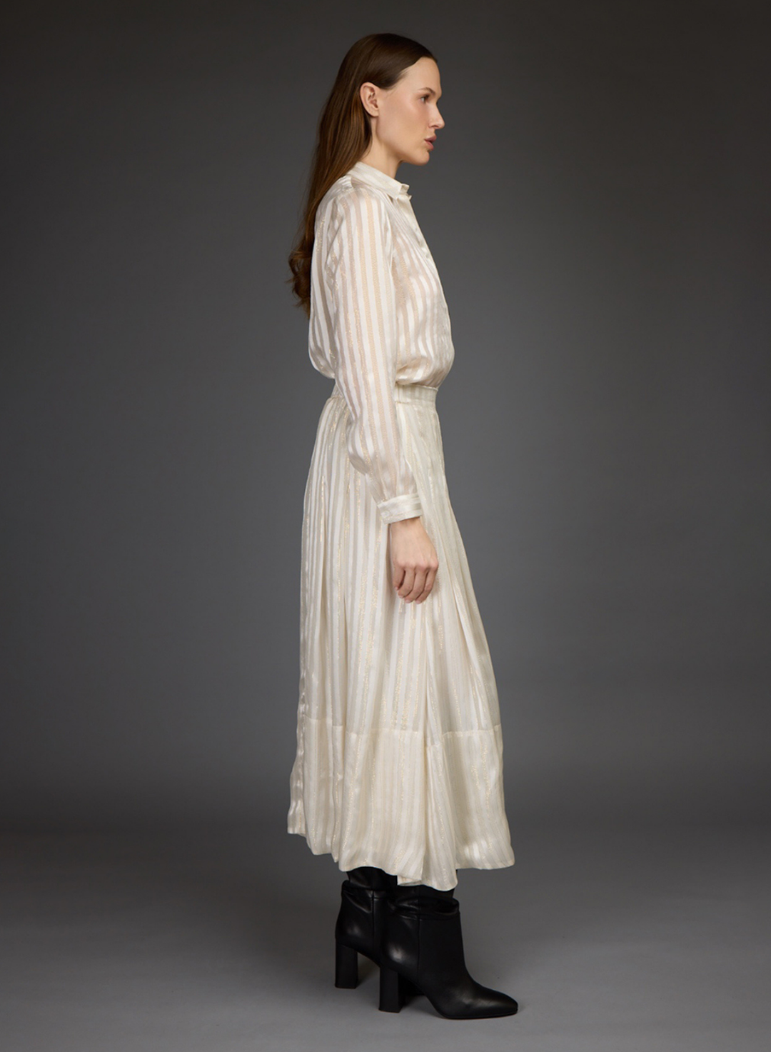A model stands against a dark gray studio background wearing a cream-colored, long-sleeved blouse and skirt set with black leather boots.