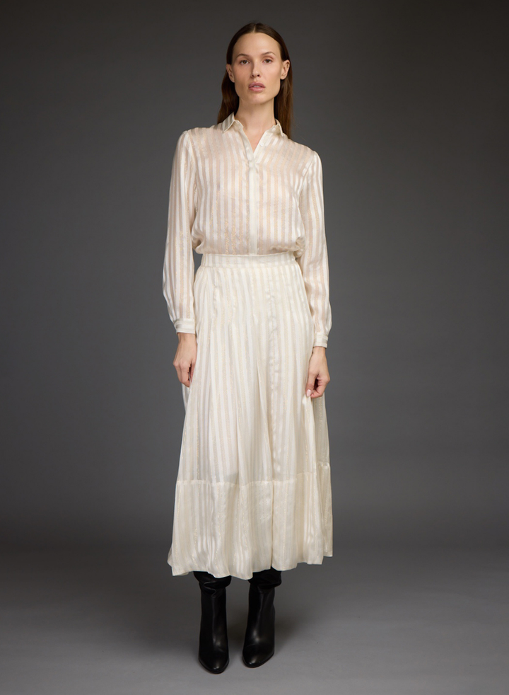 A model stands against a dark gray studio background wearing a cream-colored, long-sleeved blouse and skirt set with black leather boots.