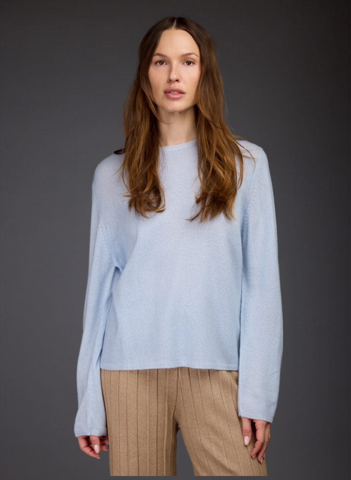 A woman modeling a light blue long-sleeve knit sweater with a crew neck, paired with beige ribbed trousers, against a dark gray studio background.