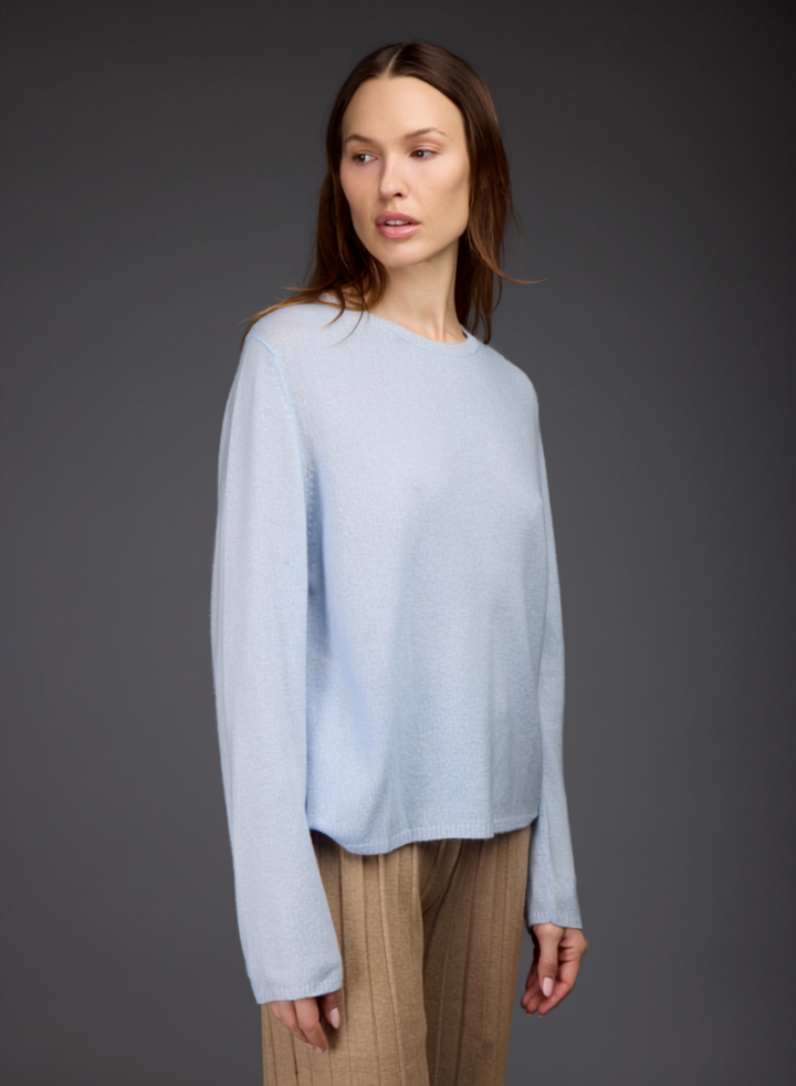A woman modeling a light blue long-sleeve knit sweater with a crew neck, paired with beige pleated trousers, against a dark gray studio background.