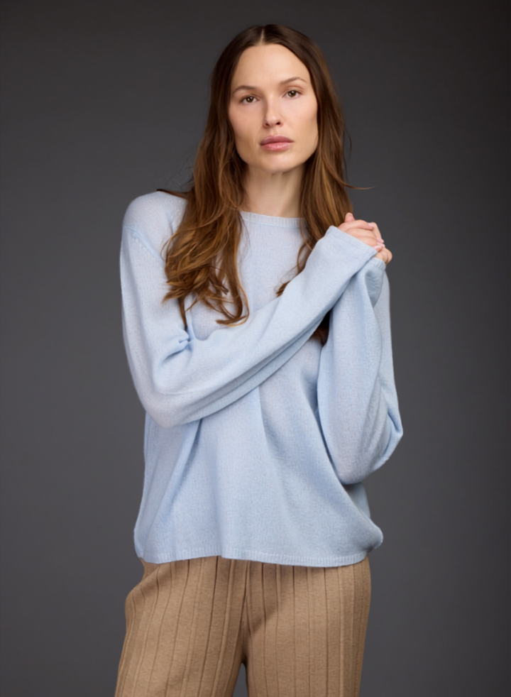 A woman modeling a light blue knit sweater with long wide sleeves and a crew neckline, paired with beige pleated trousers, against a dark gray studio background.
