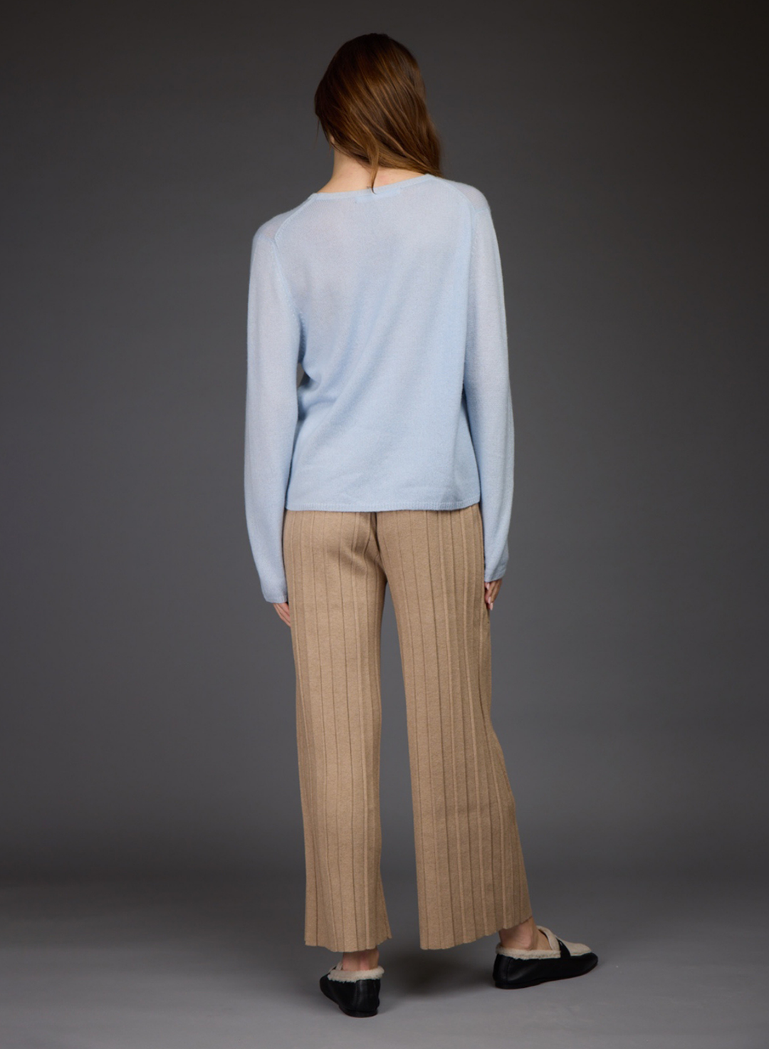 A woman shown from the back wearing a light blue long-sleeve top and beige wide-leg pleated trousers, standing against a dark gray studio background.