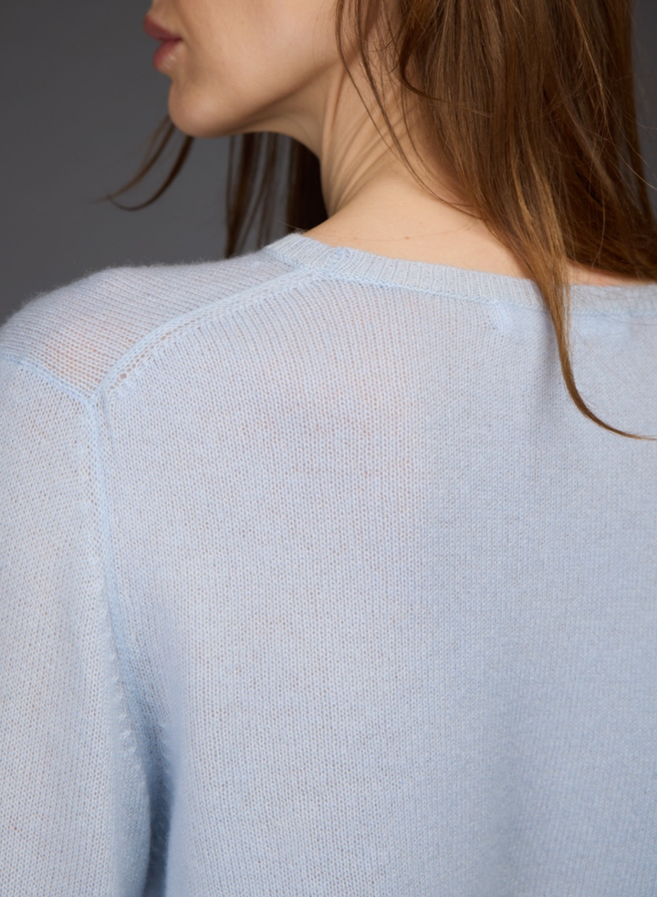 Close-up back view of a woman wearing a light blue knit sweater, showing the fine knit texture and shoulder seam against a dark gray background.