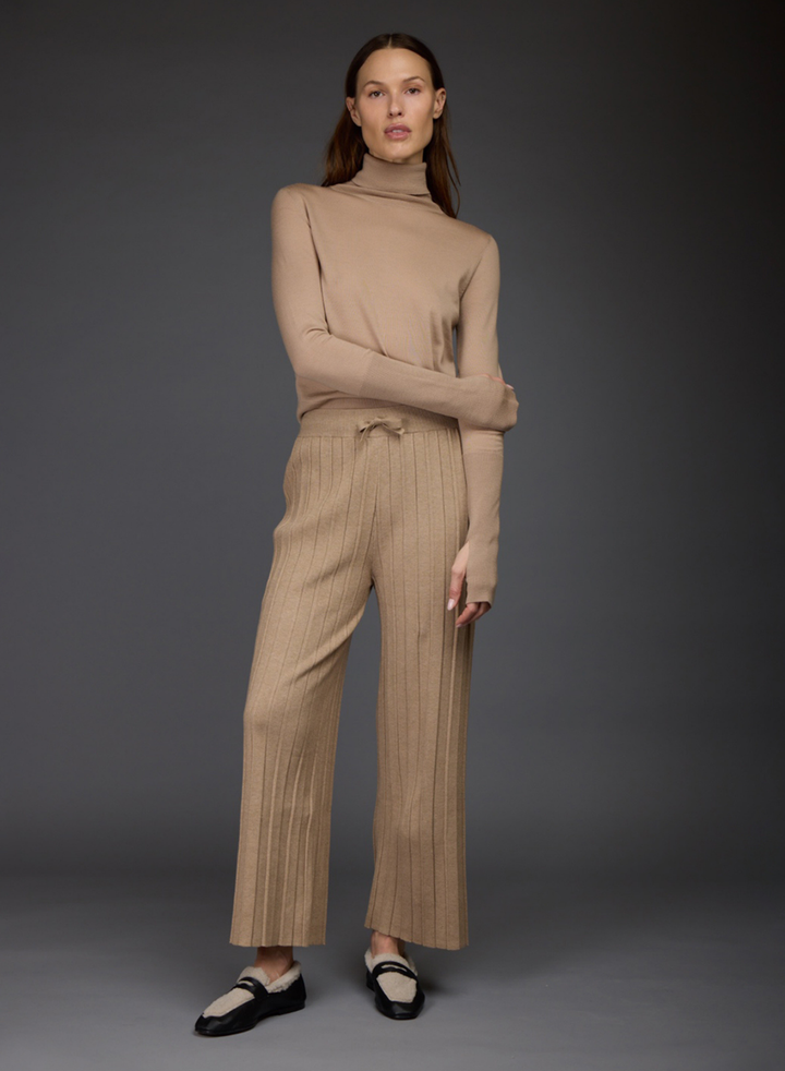 A model stands against a dark gray studio background wearing a beige ribbed turtleneck sweater by Love Binetti and matching wide-leg drawstring pants paired with black and white loafers.