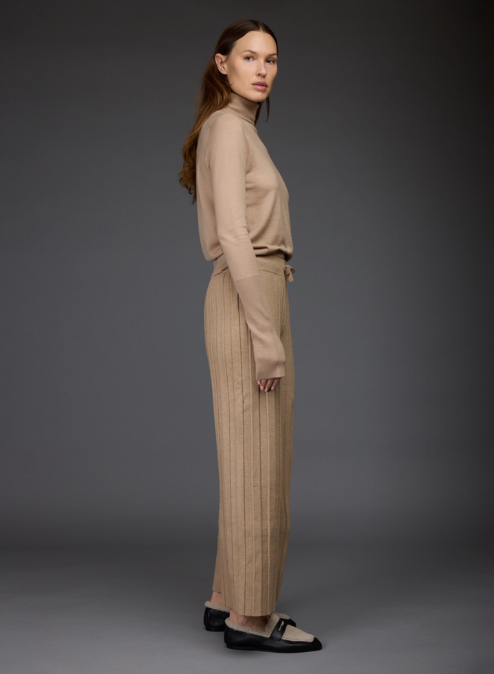 A model shot from the side against a dark gray studio background wearing a beige ribbed turtleneck sweater by Love Binetti and matching wide-leg drawstring pants paired with black and white loafers.