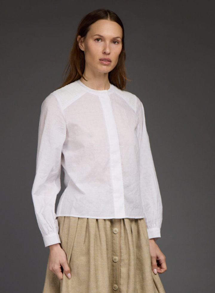 A woman modeling a white long-sleeve blouse with a sheer front panel and beige button-front skirt against a dark gray studio background.